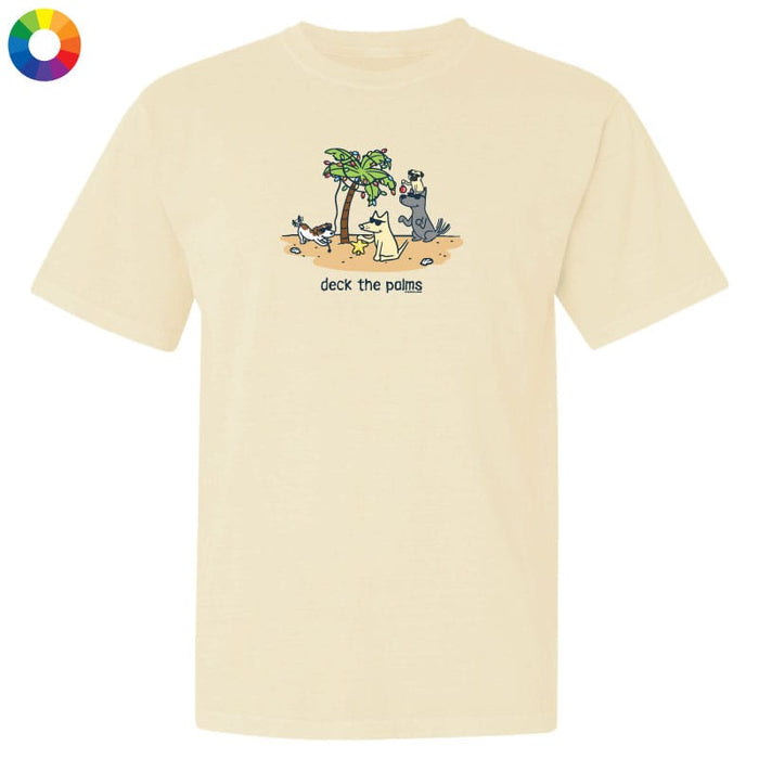 A cream-colored short-sleeve t-shirt with a colorful cartoon graphic of dogs under a palm tree and the text 'deck the palm' printed on the chest, shown on a plain white background with a color wheel icon in the top-left corner.