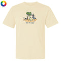 A cream-colored short-sleeve t-shirt with a colorful cartoon graphic of dogs under a palm tree and the text 'deck the palm' printed on the chest, shown on a plain white background with a color wheel icon in the top-left corner.