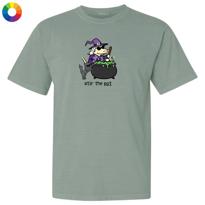 A sage green short-sleeve t-shirt with a cartoon graphic of a witch stirring a cauldron of green potion, with the text 'stir the pot' underneath. A multicolor color wheel icon is in the top-left corner.