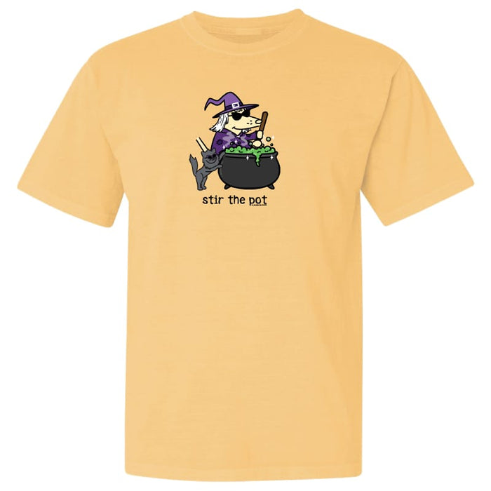 A light peach-colored short-sleeve t-shirt with a cartoon graphic of a witch sitting in a cauldron and the text 'stir the pot' printed below it.