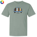 A sage green short-sleeve t-shirt with a graphic of four dogs sitting in the tailgate of a blue pickup truck, with the text 'TAIL-GATE' printed below. The t-shirt is displayed flat against a white background, with a small multicolor color wheel icon in the top-left corner.