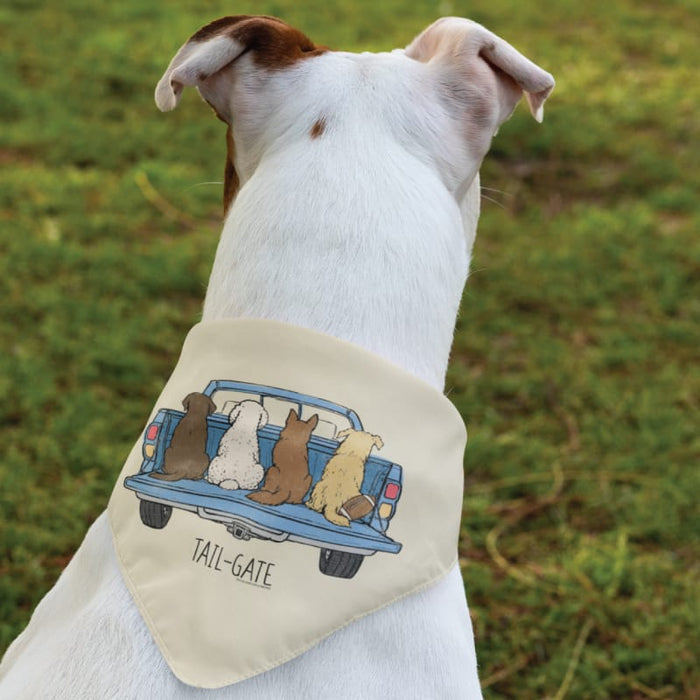 A white dog viewed from behind wearing a cream-colored bandana with a cartoon illustration of dogs sitting in the back of a blue pickup truck and the text 'TAIL-GATE'. The dog is outdoors on a grassy lawn.