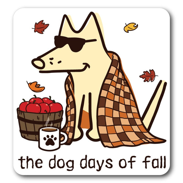 A square coaster with rounded corners featuring a cartoon dog wearing sunglasses and a brown and beige checkered blanket, sitting next to a basket of red apples and a paw-print mug. The text 'the dog days of fall' is printed at the bottom, with small autumn leaves floating around on a white background.