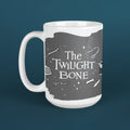 A white ceramic mug featuring a dark gray graphic print with the text 'The Twilight Bone' and small white illustrations, shown against a dark teal background.