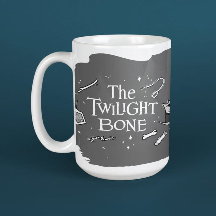 A white ceramic mug featuring a dark gray graphic print with the text 'The Twilight Bone' and small white illustrations, shown against a dark teal background.