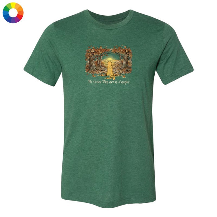A heather green short-sleeve crewneck t-shirt with a central graphic of a yellow dog running through a forest with the text 'The cows they see a dog'. A small multicolor color wheel icon is in the top-left corner, all on a solid white background.